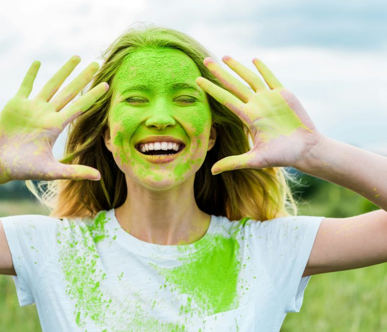 cheerful woman with closed eyes and green holi paint on hands gesturing and smiling outdoors